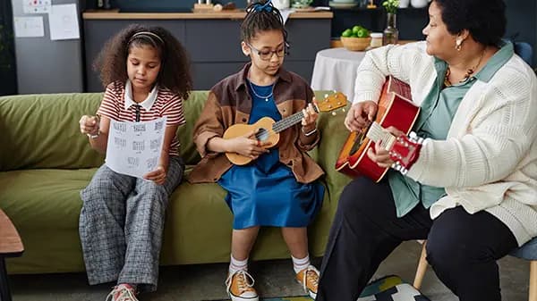 Children learning guitar alongside an adult, sitting on a couch together.